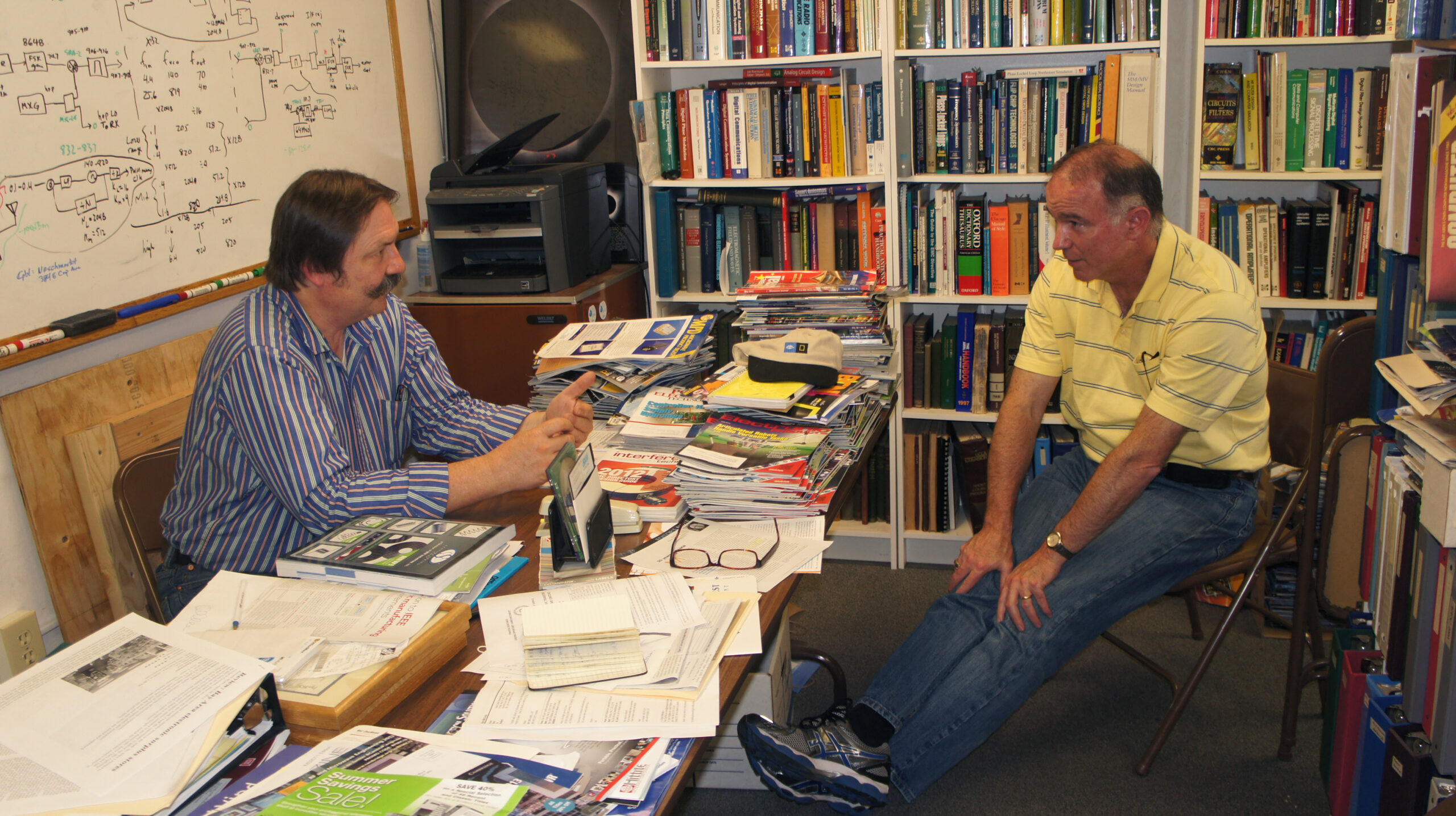 doug (wearing a yellow shirt) and earl (wearing a blue button down) sit down in an office, talking.